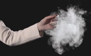 View of an arm with the hand touching a puff of smoke in a space with a dark background