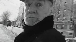 Black and white footage of George Carlin dressed in a black coat, looking surprised, featured in HBO's doc, George Carlin's American Dream.