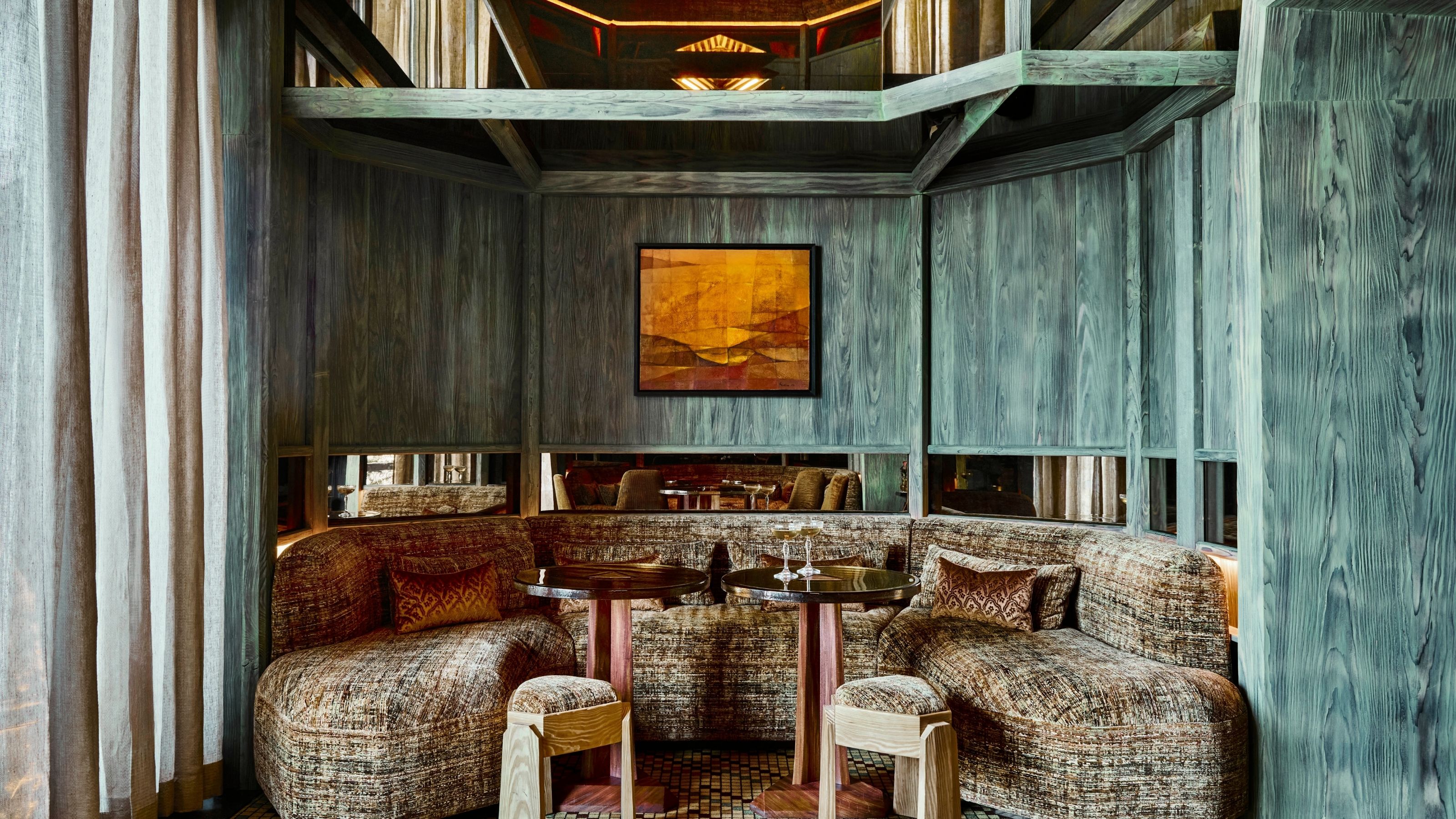 A seating booth carved from blue-painted wood, enveloping upholstered banquettes, wood and textile stools, and a mirrored panel breaking the wall into two is captured frontally.