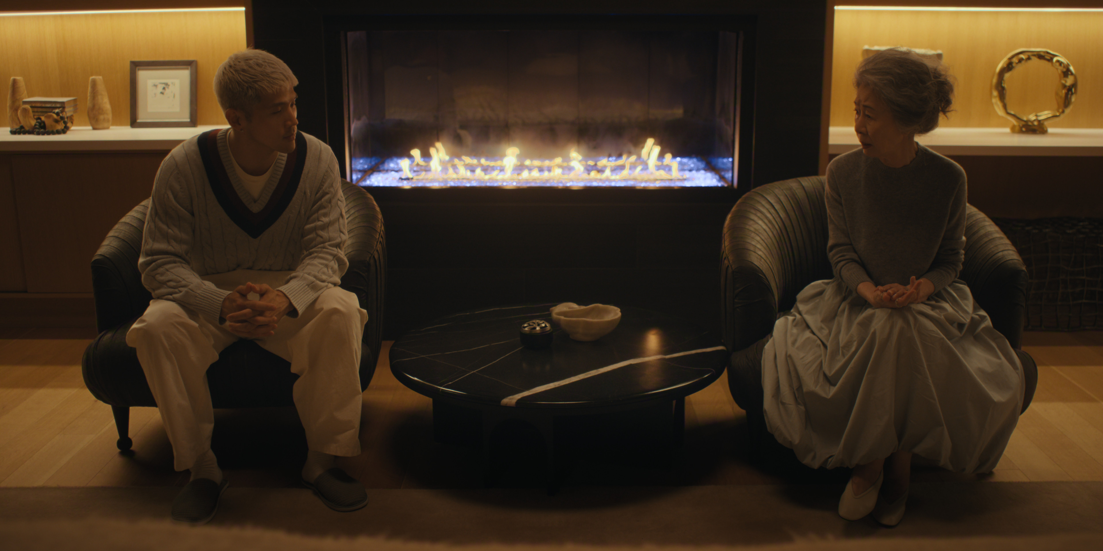 A young man (Matthew Kim as Woosh) and an older woman (Youn Yuh-jung as Chairwoman Park) sit in chairs on either side of a fireplace, in episode 206 of 'Beef.'
