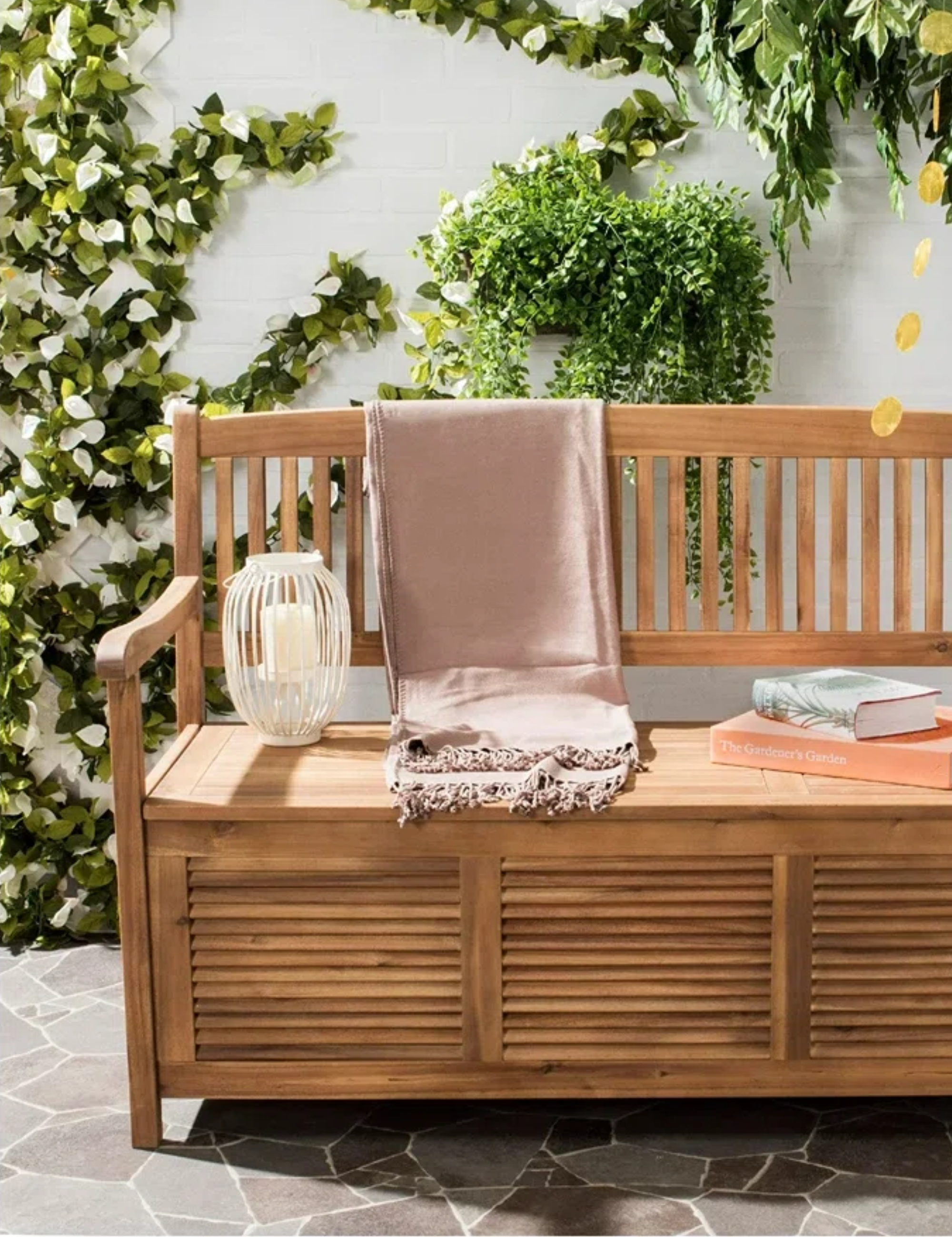 Wooden outdoor storage bench styled atop of a stone patio area alongside a lantern, throw blanket, and books