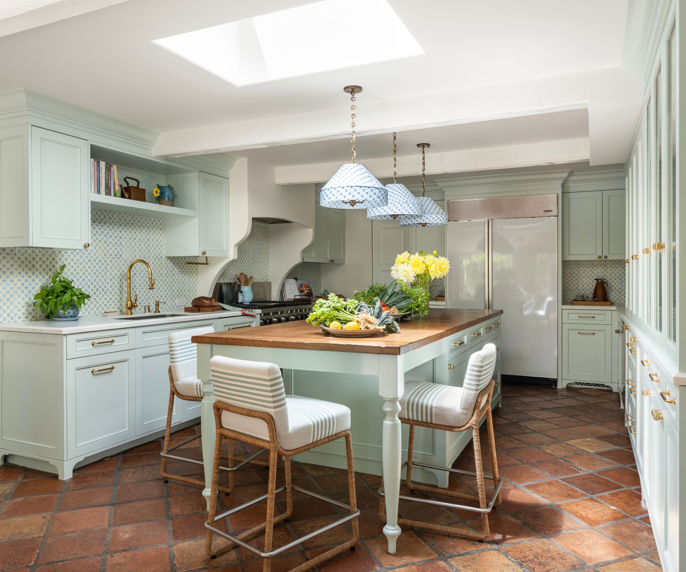 a blue/green kitchen with terracotta floor tiles, a large island with a wooden countertop, and patterned pendant light shades