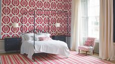 A red and white bedroom with patterned wallpaper and rug, and large black four poster bed with white bedding, black bedside tables, a white and red upholstered chair, and white curtains over a large window.