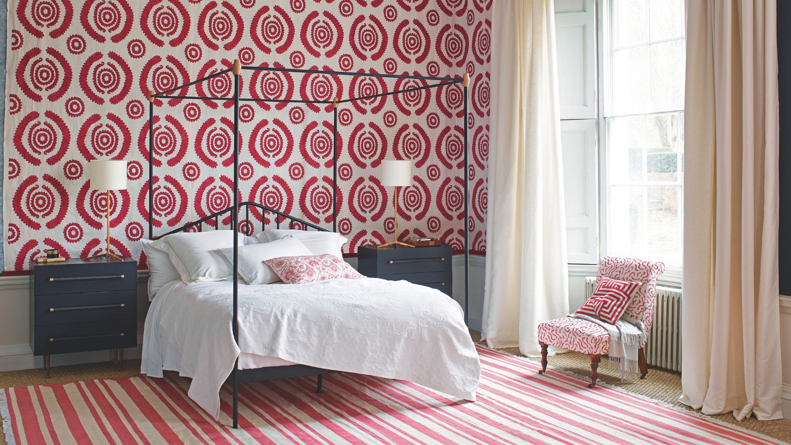 A red and white bedroom with patterned wallpaper and rug, and large black four poster bed with white bedding, black bedside tables, a white and red upholstered chair, and white curtains over a large window.