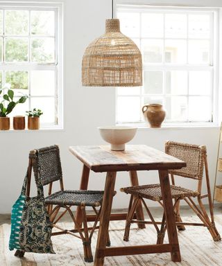 A boho dining room with a white wall with two large windows, a rattan pendant light, a dark wooden table with a white bowl, a blue rattan chair with a bag on to the left and a brown rattan chair to the right