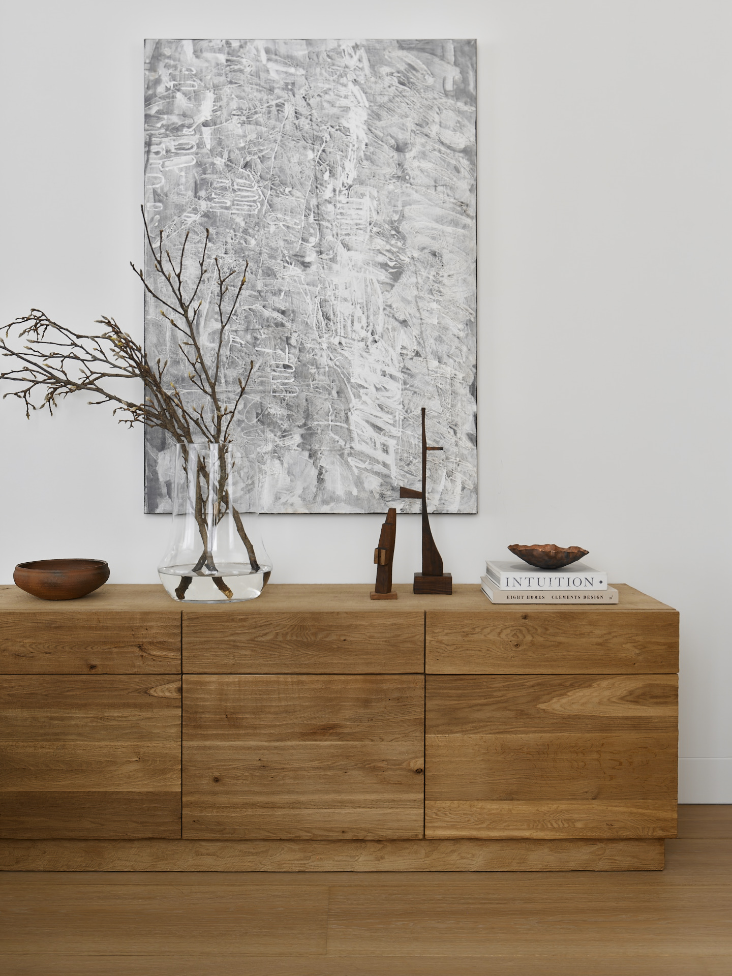 A wooden side table in an entryway with art work above and accessories on top, including a vase and plants