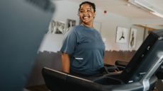 A woman in a sports top stands on a treadmill and smiles at something off-camera.