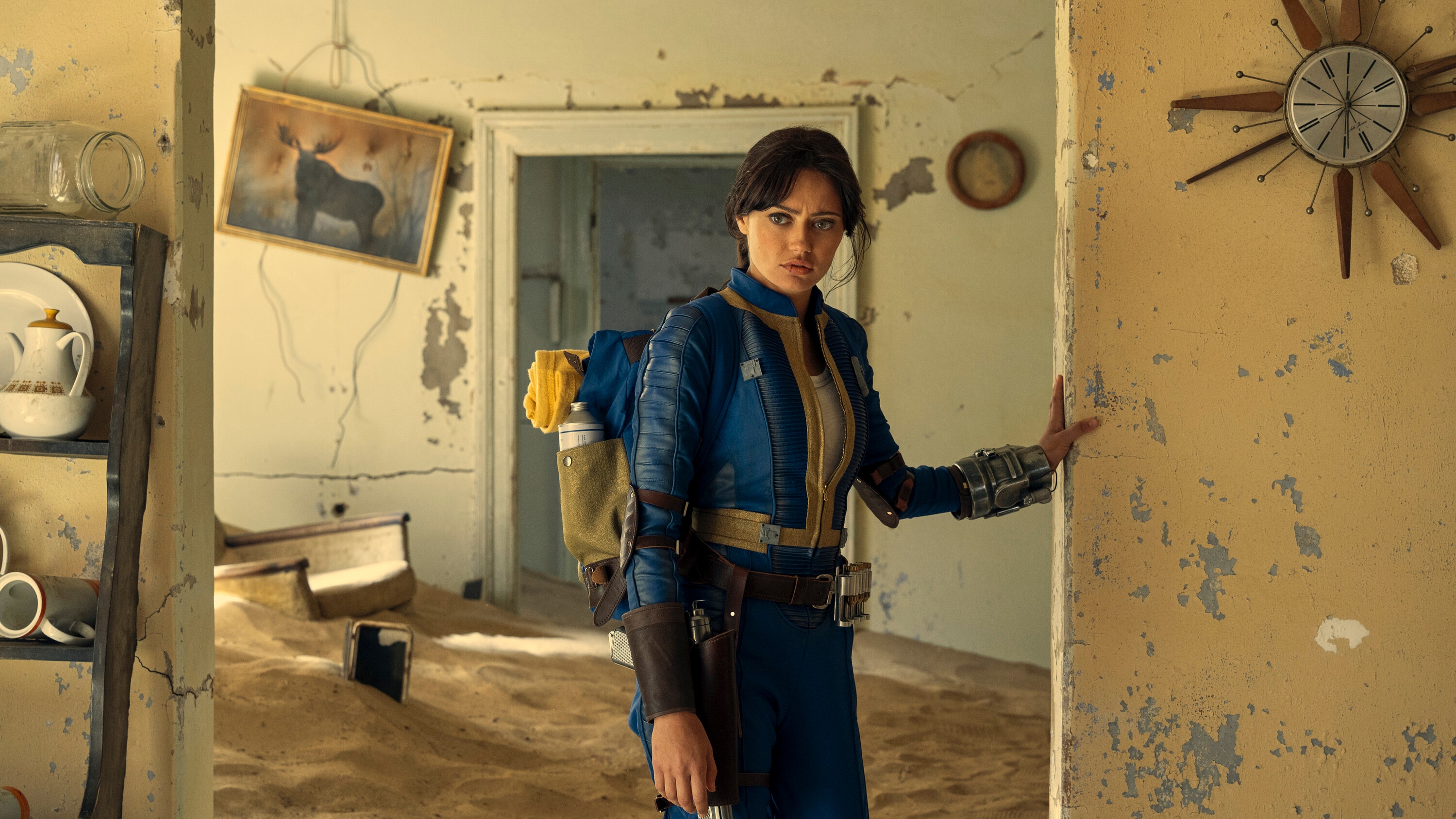 Ella Purnell como Lucy vestida con un traje azul y amarillo y mirando alrededor de una casa abandonada en Fallout.