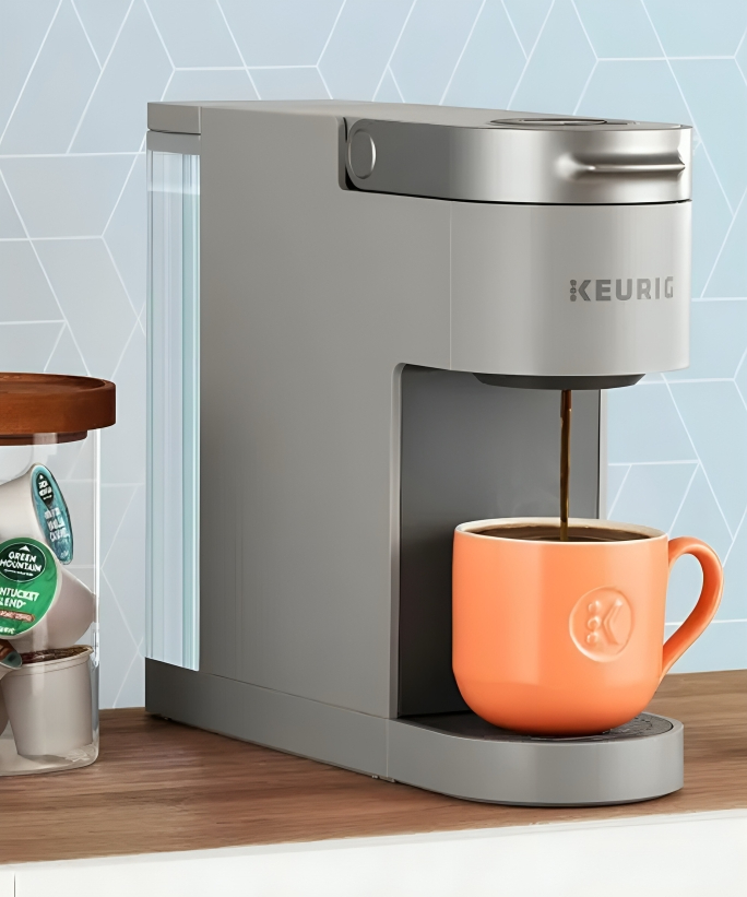 A grey slim coffee maker with an orange mug underneath the spout as coffee streams into it. On a wooden countertop with a blue wallpaper background. Half of a jar of K-Cups visible on the left hand side.