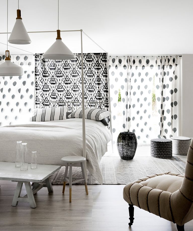 白色卧室以黑白图案床头板和透明窗帘为特色，配有一张四张海报的床，黑白家具和一把灰褐色扶手椅。