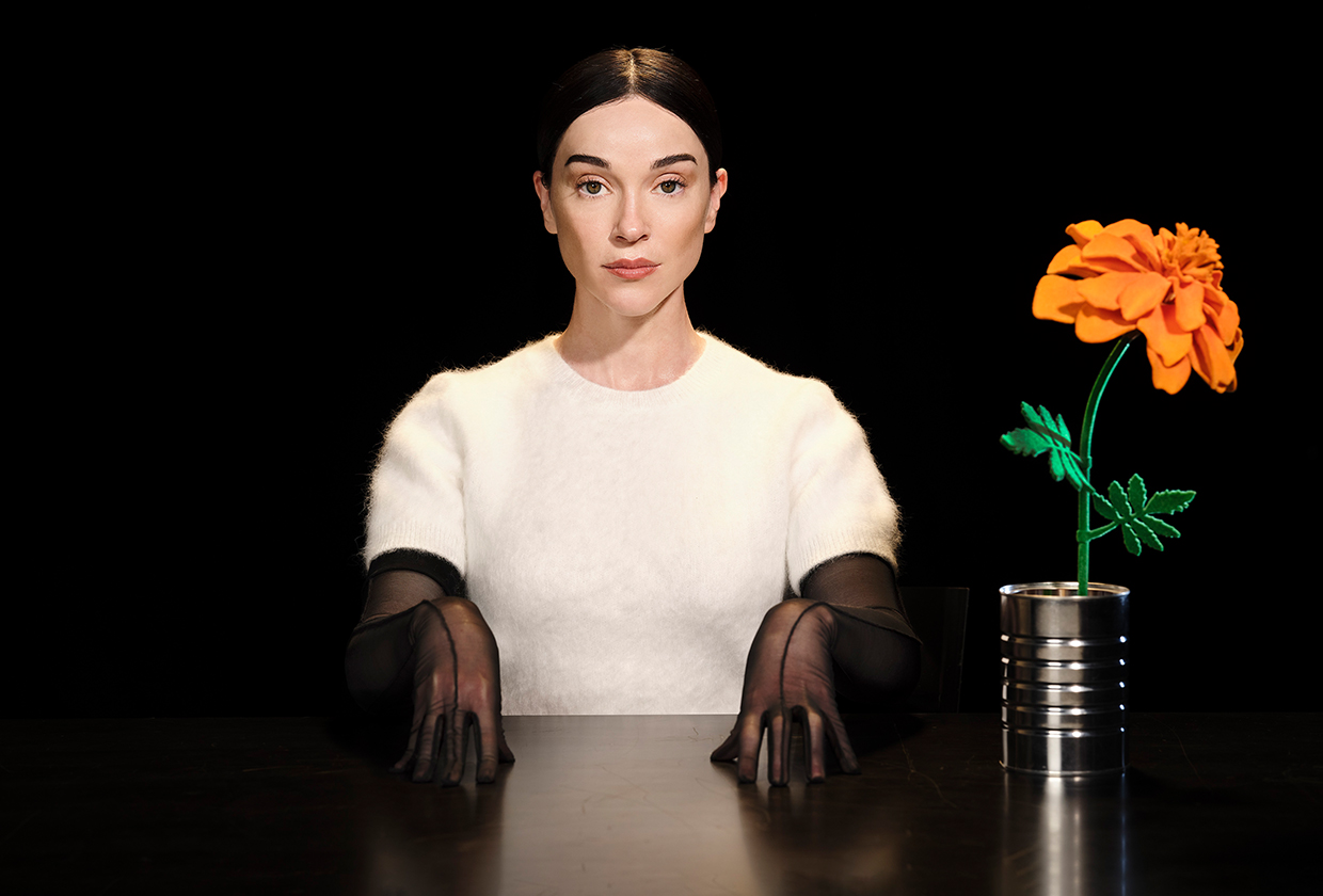 Annie Clarke, aka St. Vincent, sits at table in white short sleeve sweater and black sheer gloves next to large orange flower.