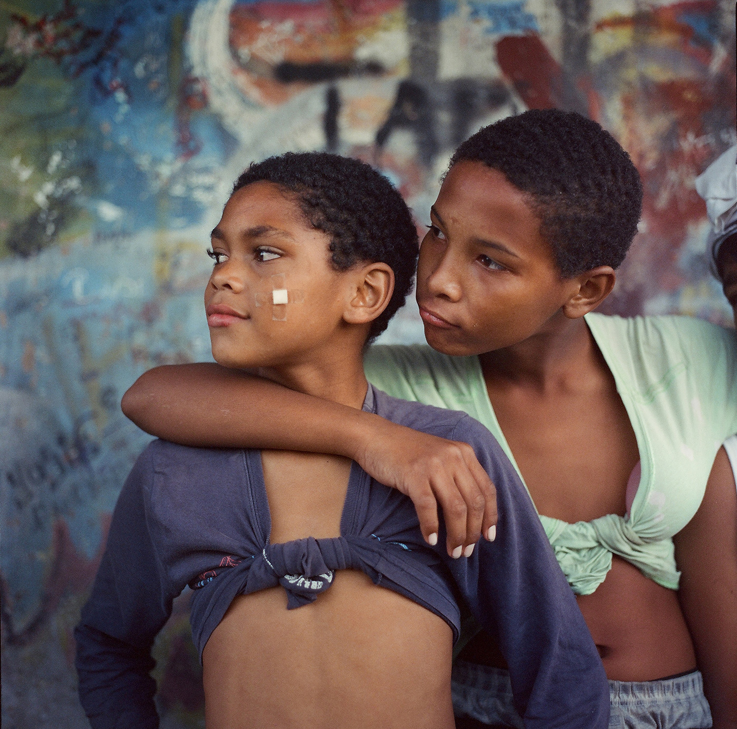 Two young boys are shown in a close-up, with one boy in a light green top wrapping his arm around the shoulders of the other boy, who has a small bandage on his cheek.