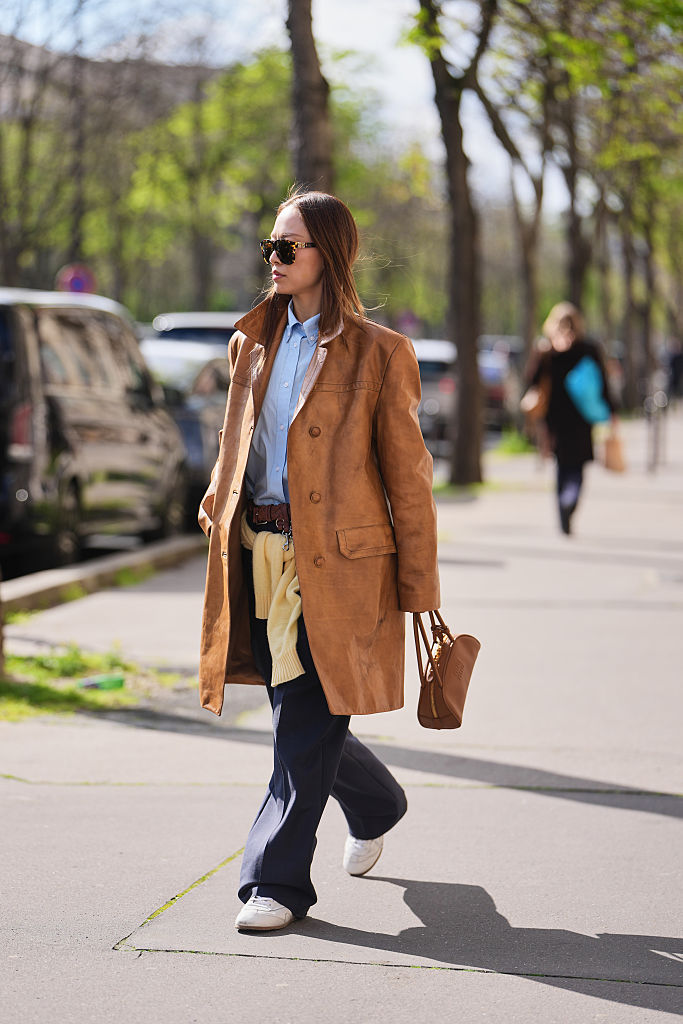 Niki Wu Jie wears black sunglasses, a brown suede double breasted coat, a light blue cotton button down shirt, a brown braided leather belt, dark blue wide leg trousers, a pale yellow pullover, a brown leather bag from Miu Miu, white leather sneakers shoes, outside Miu Miu, during Paris Fashion Week