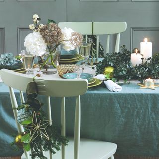 A dining table set for a Christmas dinner in a light blue-green colour scheme with a vase of flowers and a eucalyptus wreath in the middle