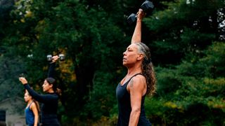Woman in park holding dumbbell overhead, more women are exercising with dumbbells in the background