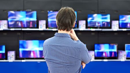 A man thoughtfully looks at LCD TVs in a large store with his back to the camera