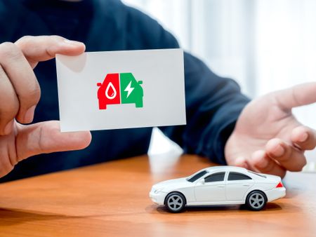 Electric energy and fuel engine, plug-in hybrid electric vehicles, on white business card in broker or agent hand near car toy on wood table.
