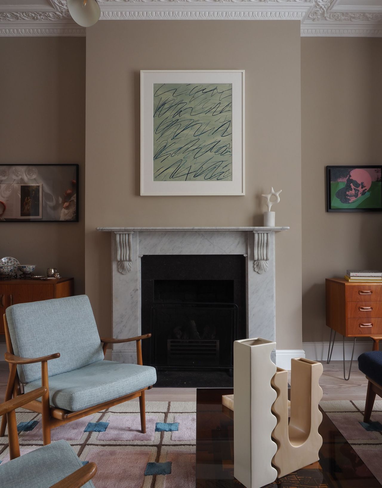 A modern living room with interconnecting sculptural vases and a blue cushioned and wood framed armchair on top of a patterned rug in front of the fireplace with an artwork on top and either side