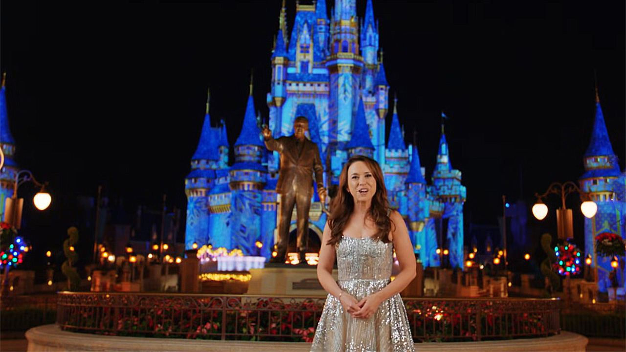 Lacey Chabert stands in front of the famous castle lit up at night and statue of Walt Disney at Disney World.