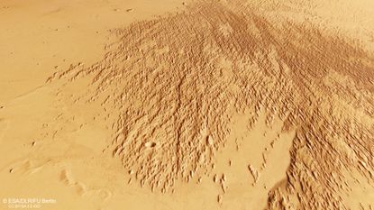 A section of Mars&rsquo;s surface showing a broad area covered with dense, elongated ridges and grooves that fan out diagonally across the image. The terrain is rough and textured in this region, contrasting with the smoother, lighter-coloured surrounding plains. The overall colour is a mix of tan and reddish-brown tones.