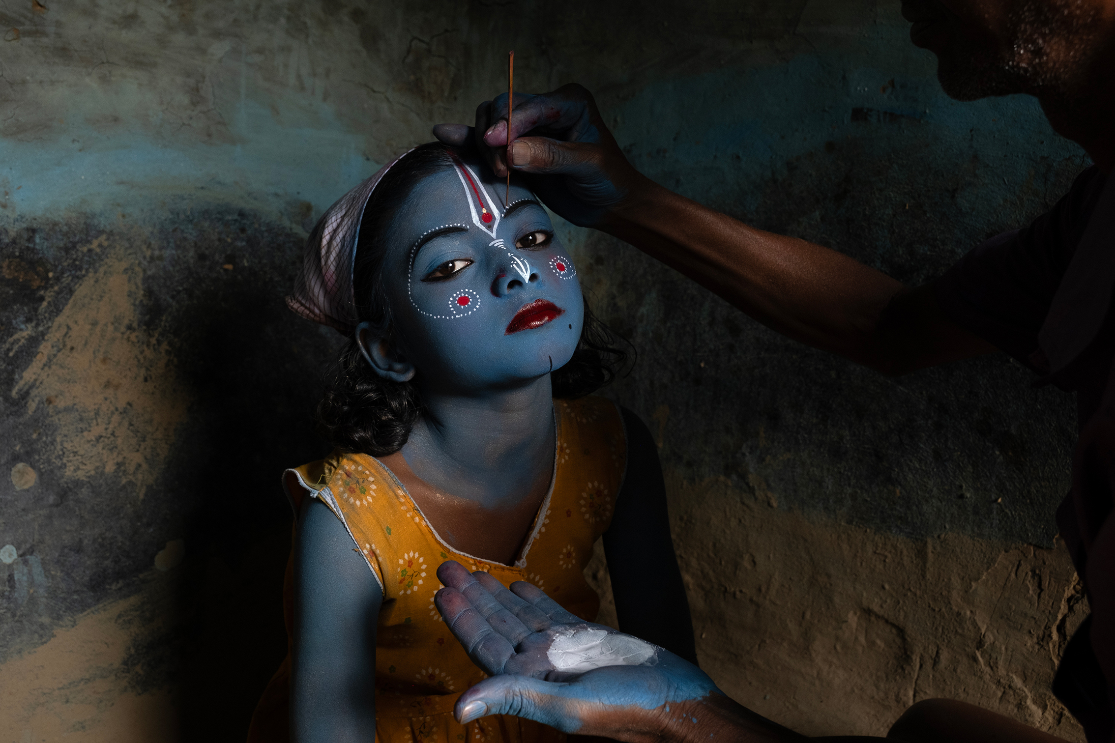 A child with blue skin sits calmly as an adult applies paint to their face in a dimly lit room