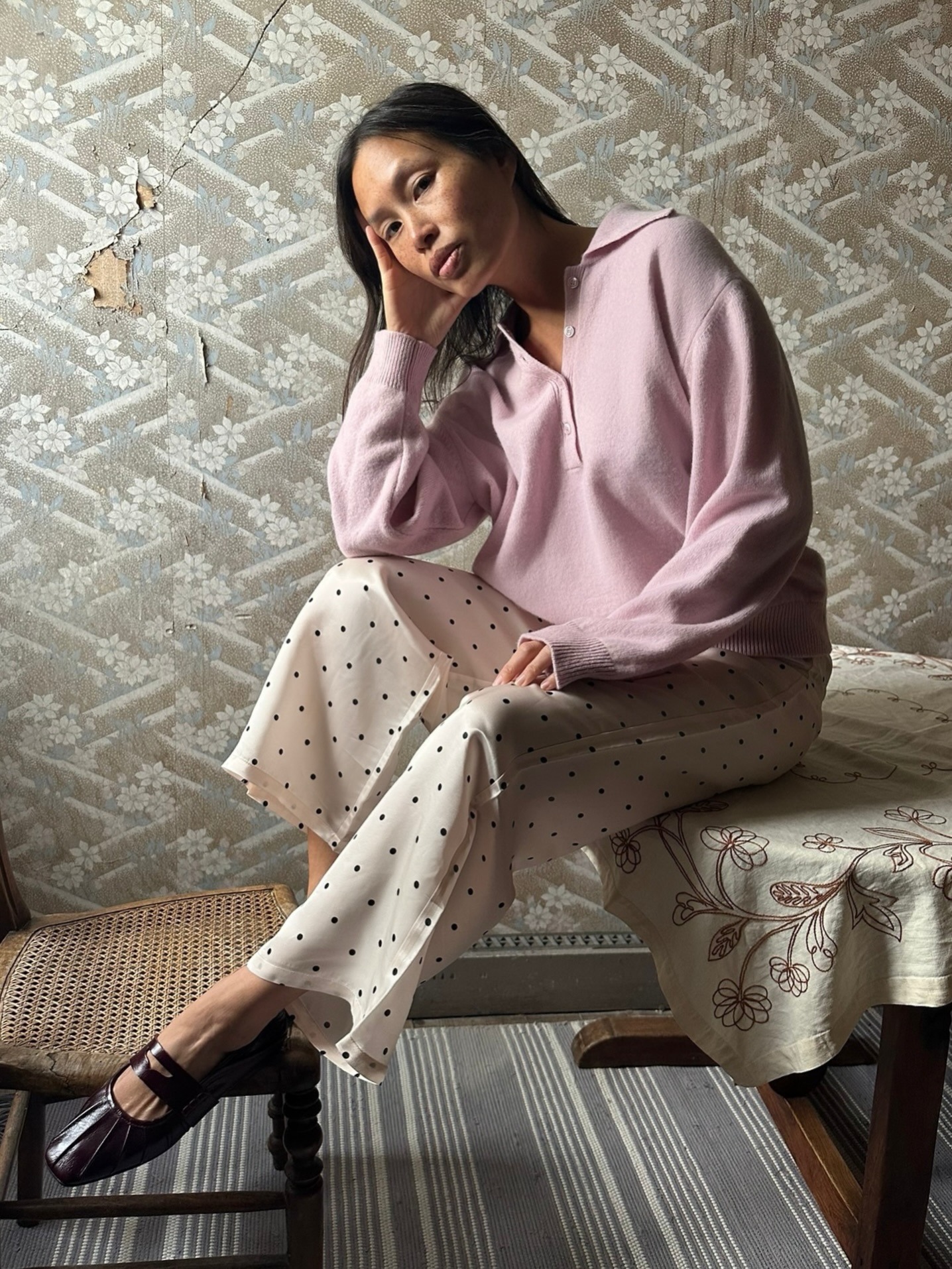 French fashion influencer Anne-Victoire Lefevre leaning against a baby blue wall and on ornate blue tiles wearing a stone pendant cord choker necklace, brown off-the-shoulder long-sleeve maxi dress, and black knit lace-up flats