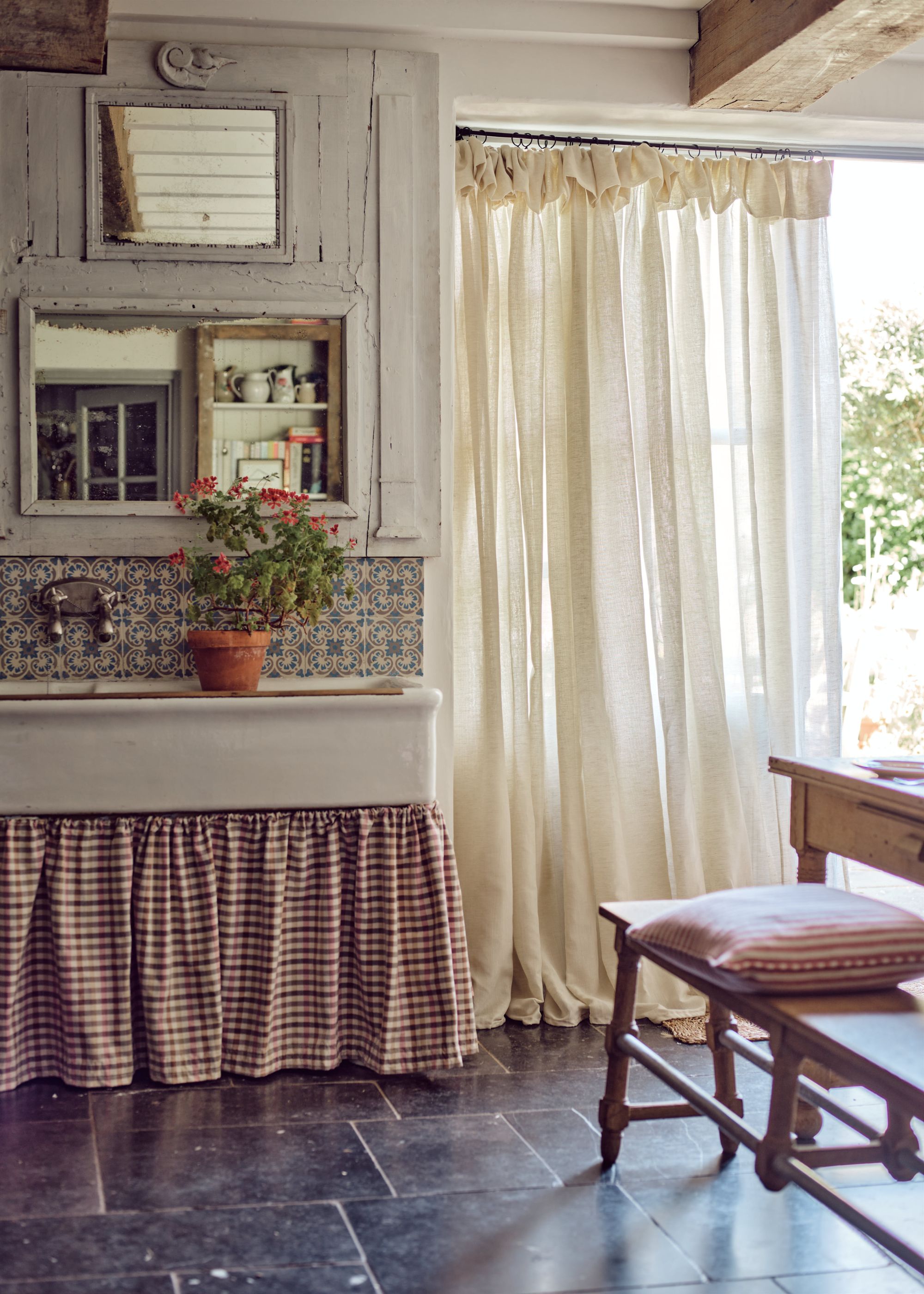 A rustic room with an external door with a neutral linen curtain, and a butler's sink in the corner with a gingham curtain.