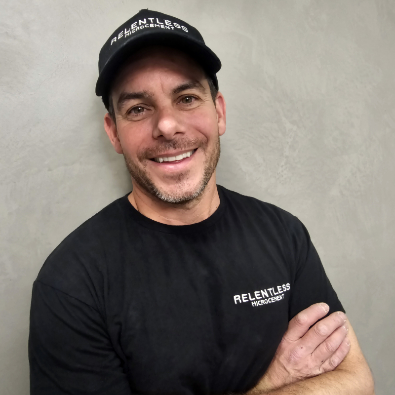 male wearing black baseball cap and black tshirt with arms folded smiling at camera