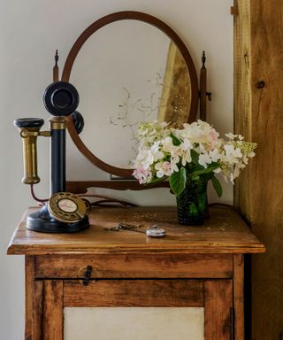 Wooden cupboard with old style telephone, vase of white hydrangeas and a mirror.