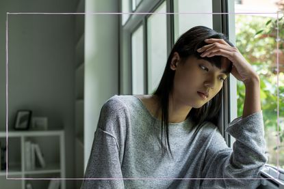 young female adult struggling with mental health looking out of window with hand on her head