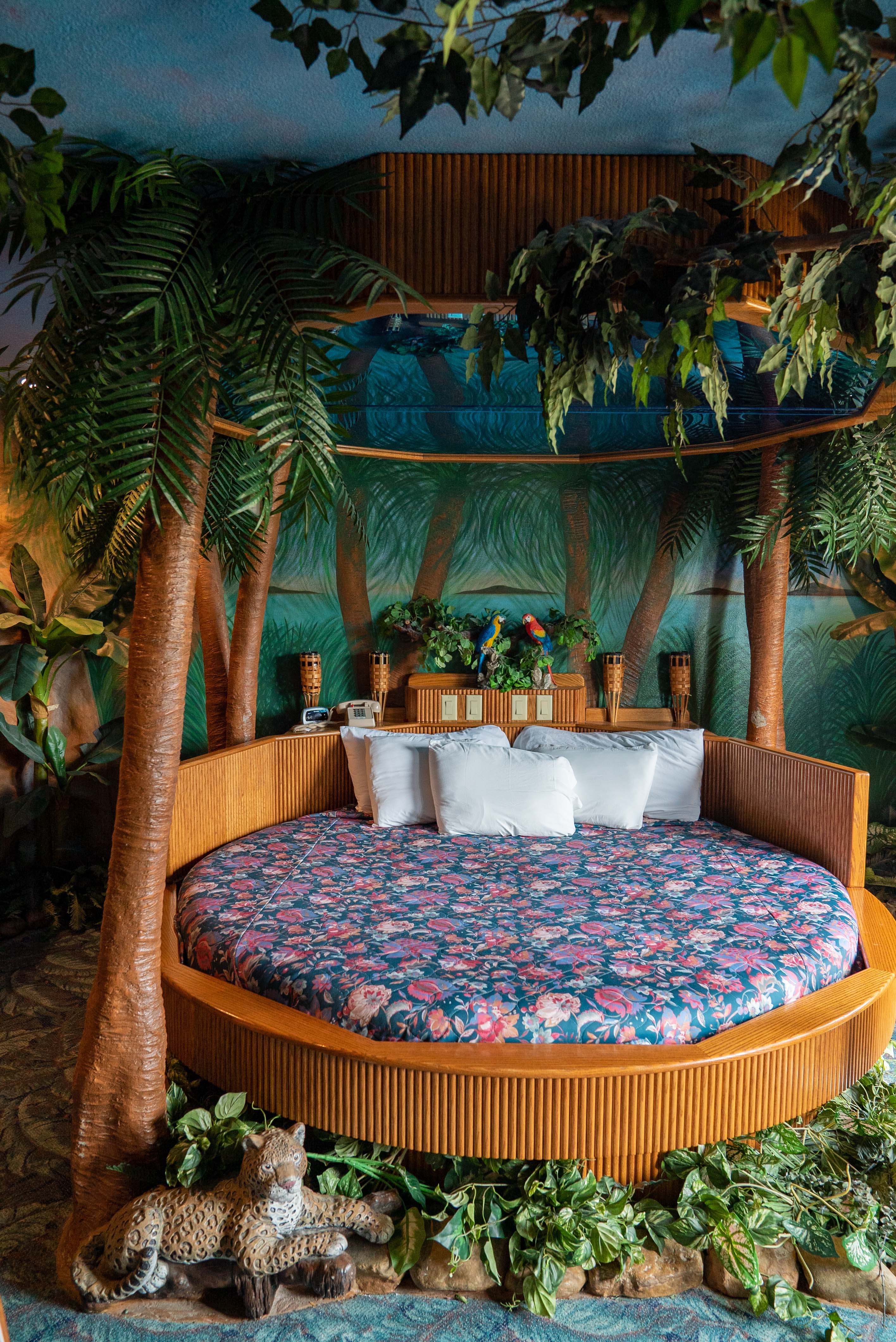 A round-shaped bedframe houses a flowery mattress in rose and blue tones and four white pillows in a tropical-themed bedroom.