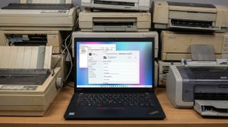 A Lenovo laptop running Windows 11 showing Printer settings with a bunch of old, dusty printers behind the laptop