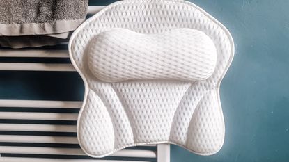 We love this ergonomic bath pillow. Here is a white mesh butterfly-shaped bath pillow hanging from a white towel rail with a gray towel next to it and a dark blue wall behind it