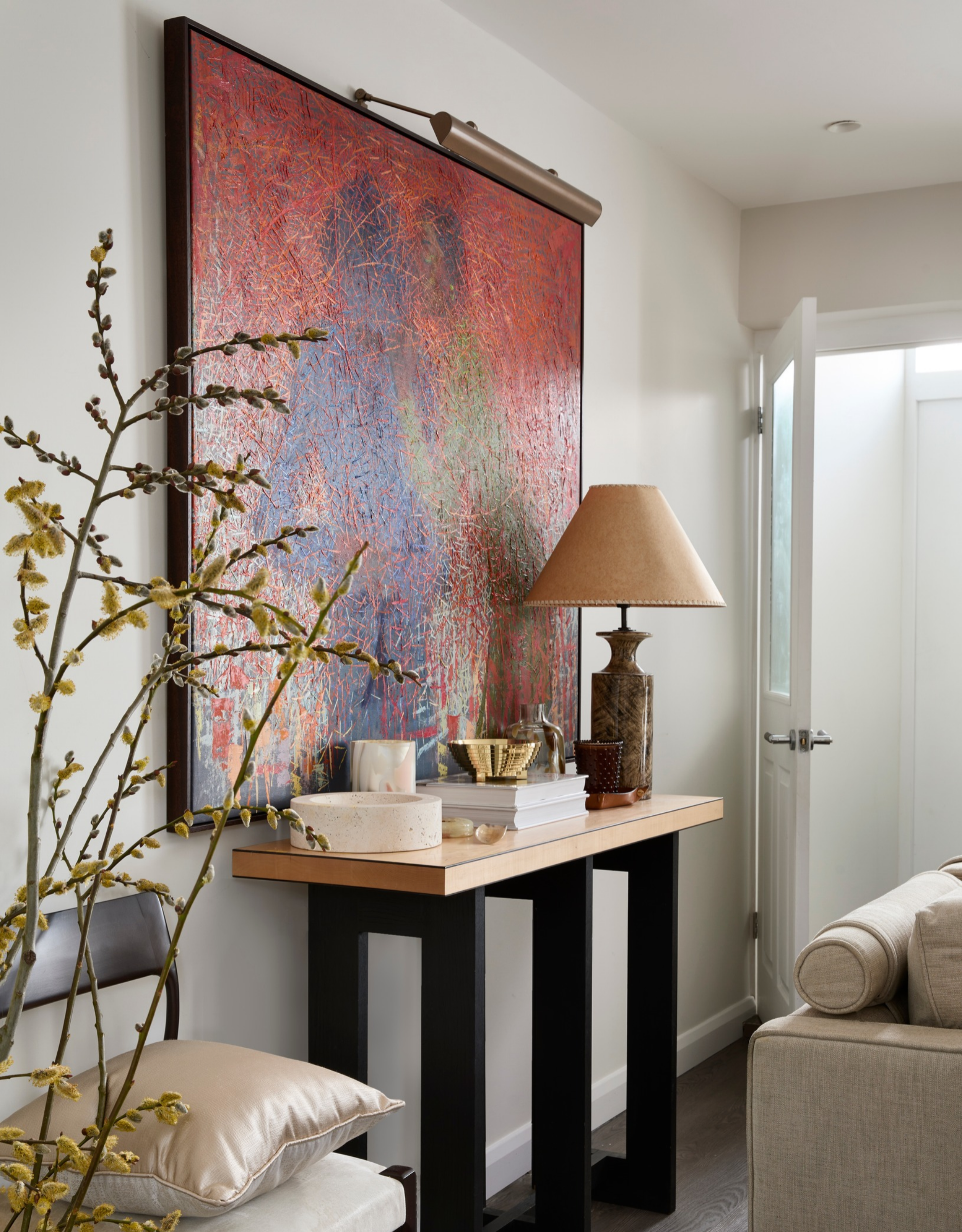 A white living room with a colorful framed artwork on the wall above a sideboard with a lamp and books