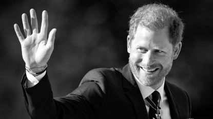 A black and white photo of Prince Harry waving and smiling wearing a suit and tie