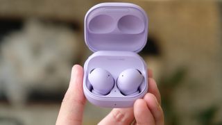 Purple Galaxy Buds Pro in their case being held by a hand so they take up the middle of the frame, they're being held in front of a blurry living room background