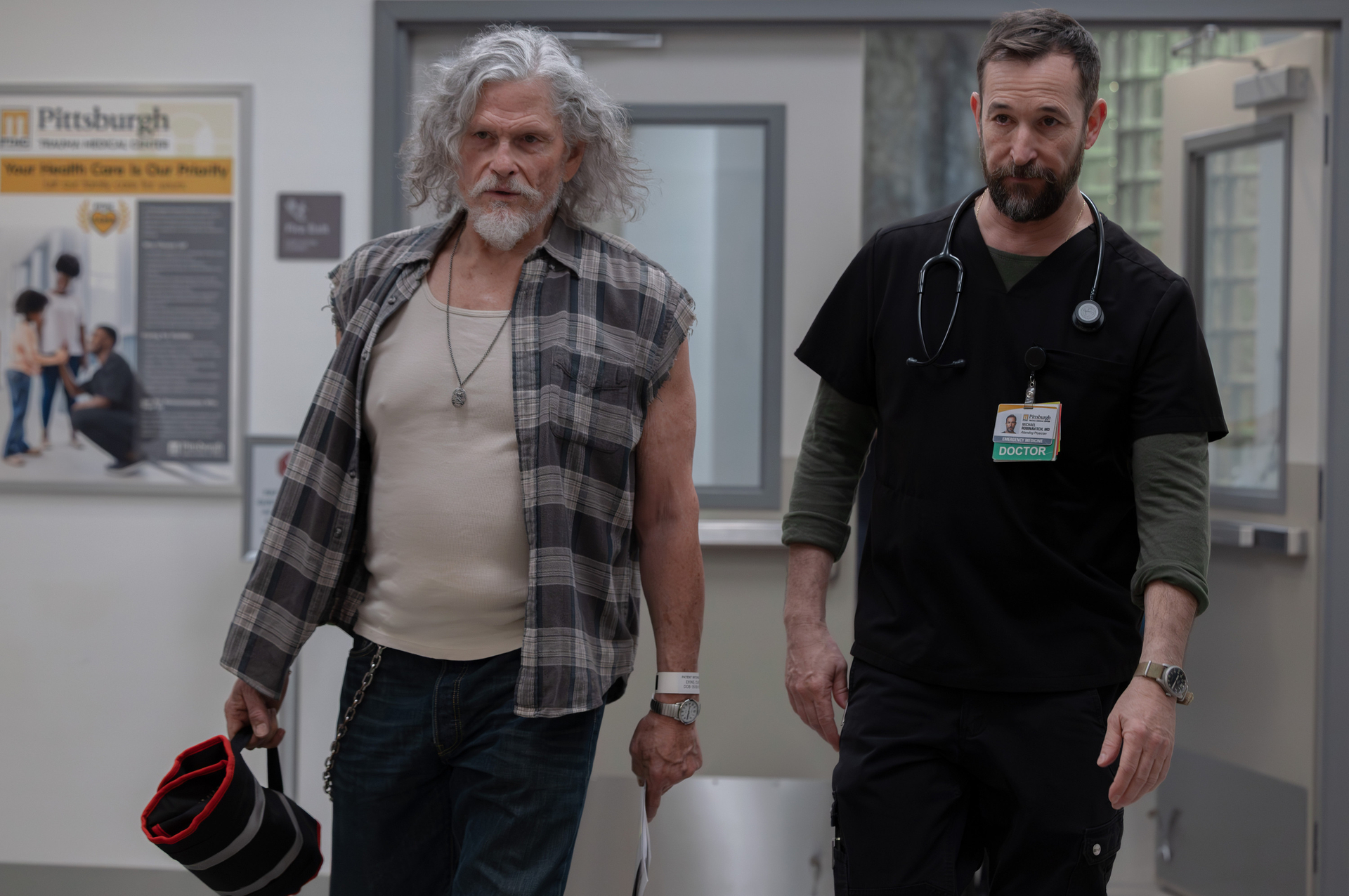 jeff kober as duke wearing a plaid shirt and noah wyle as dr robby in scrubs walking through a hospital lobby together in the pitt season 2 finale