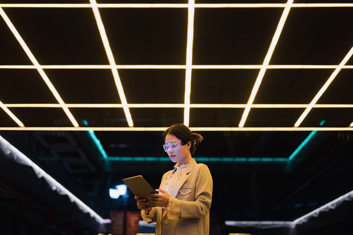 Microsoft: Woman holding an iPad in a high-tech office wearing digital glasses to denote AI assitance