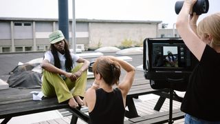 Photo shoot on rooftop with barefoot model in green pants and matching hat sitting on bench while a photographer captures her image