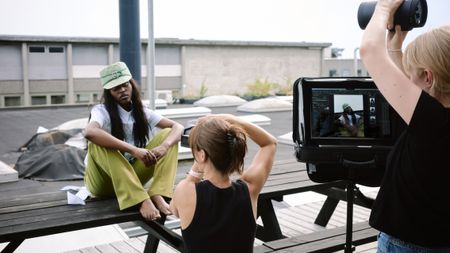 Photo shoot on rooftop with barefoot model in green pants and matching hat sitting on bench while a photographer captures her image