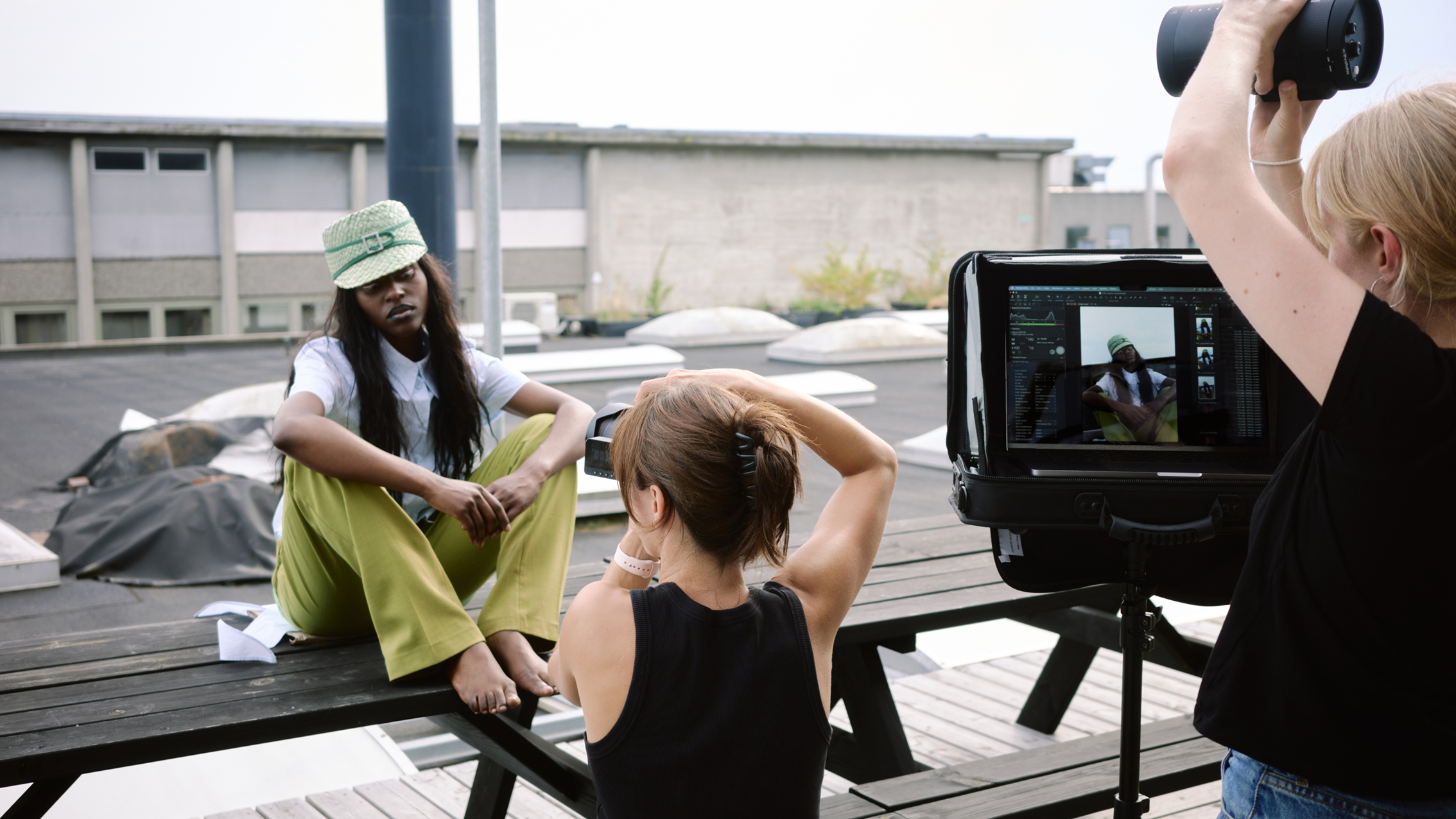 Photo shoot on rooftop with barefoot model in green pants and matching hat sitting on bench while a photographer captures her image
