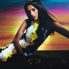 A photo of Simone Ashley sitting down in front of a colorful backdrop wearing a black, white, and yellow Missoni dress. 