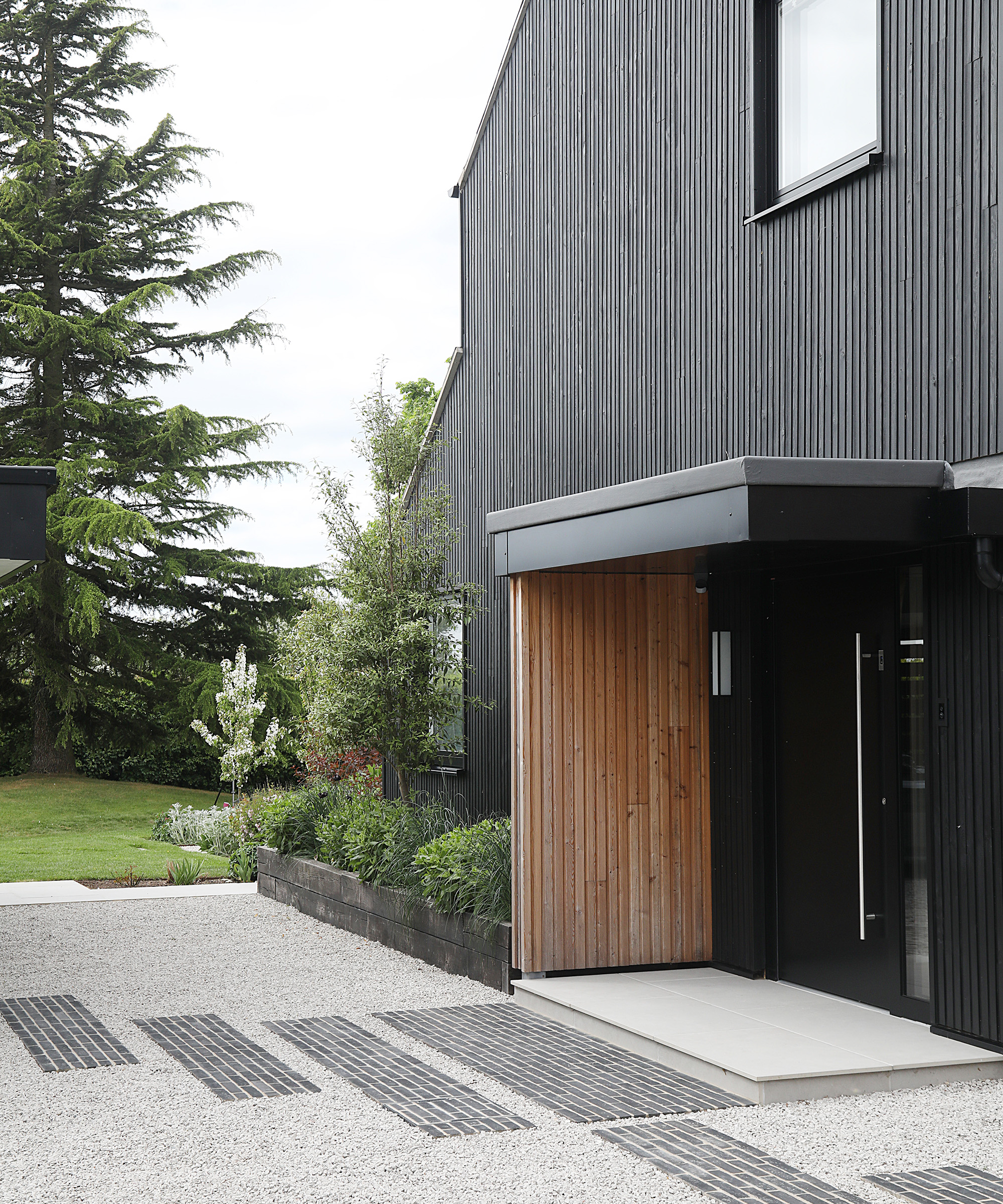A front door of a contemporary home clad in dark timber with light grey gravel leading up to the front door