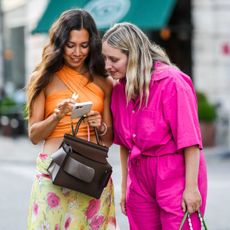 Two women on their phone searching for the best prime day beauty deals. Woman on the right in orange crop top and floral skirt smiling down at her phone. Blonde woman on the left in pink jumpsuit looking over at her phone