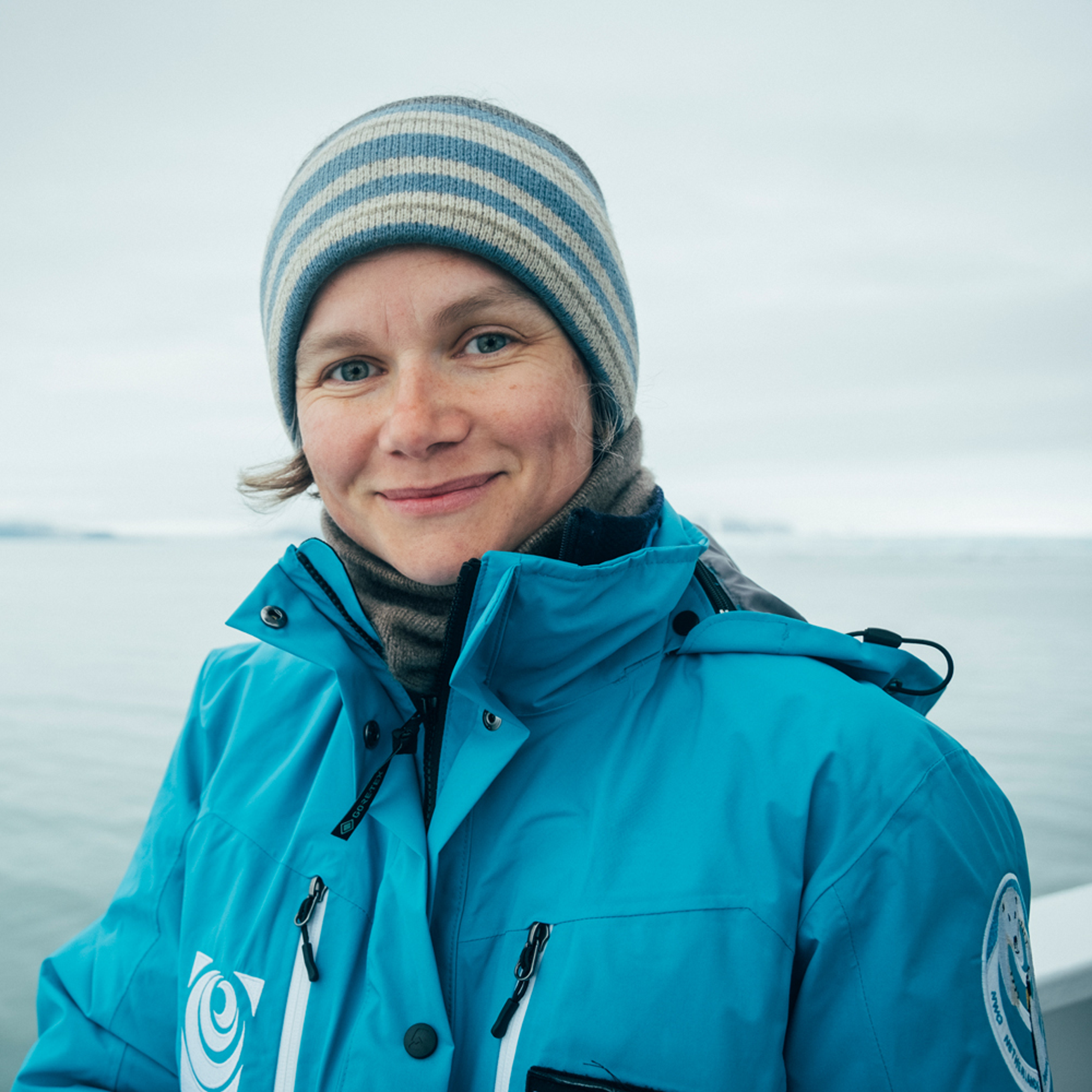 A woman wearing a blue stripped beanie and large blue winter jacket smiles at the camera