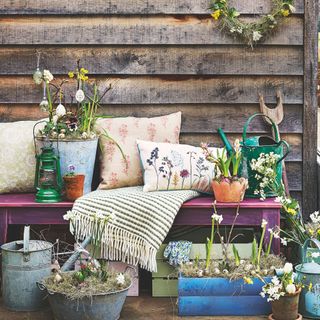 Wooden bench in front of a shed covered in cushions, throws, and other gardening accessories