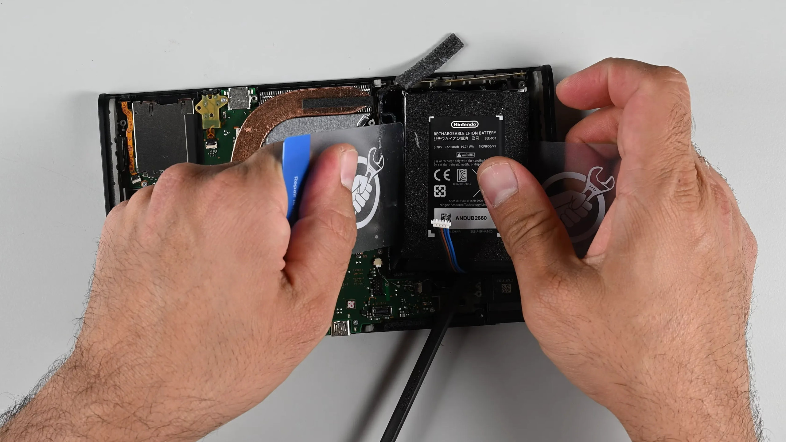 A screenshot of iFixit's Nintendo Switch 2 teardown video, showing the presenter struggling to remove the internal battery