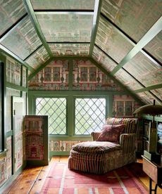 Colorful attic room with green painted trim, colorful wallpaper, striped chaise longue with patterned cushion, wooden floor with rug, bookshelf to the right.