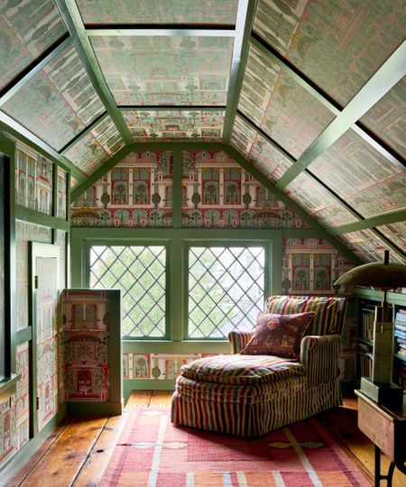 Colorful attic room with green painted trim, colorful wallpaper, striped chaise longue with patterned cushion, wooden floor with rug, bookshelf to the right.