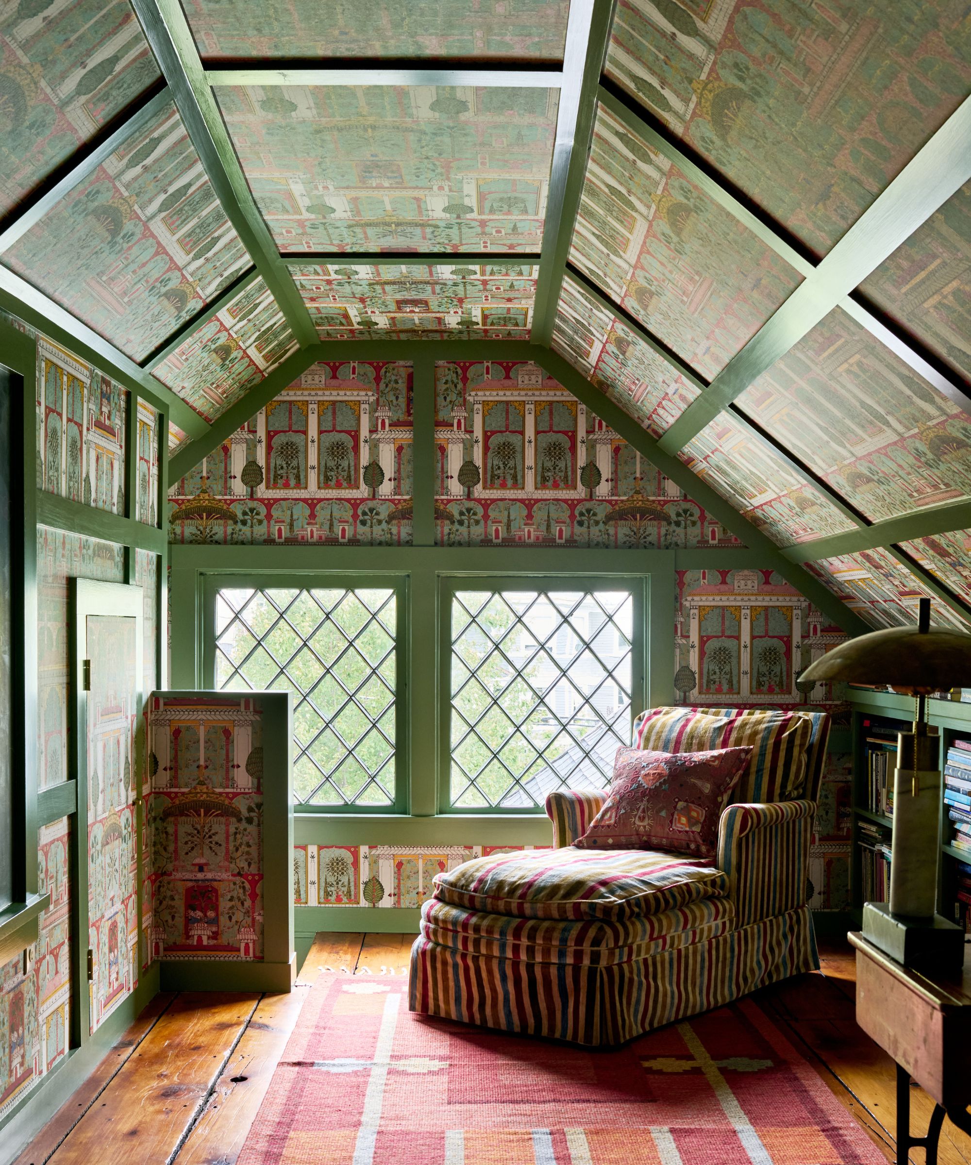 Colorful attic room with green painted trim, colorful wallpaper, striped chaise longue with patterned cushion, wooden floor with rug, bookshelf to the right.